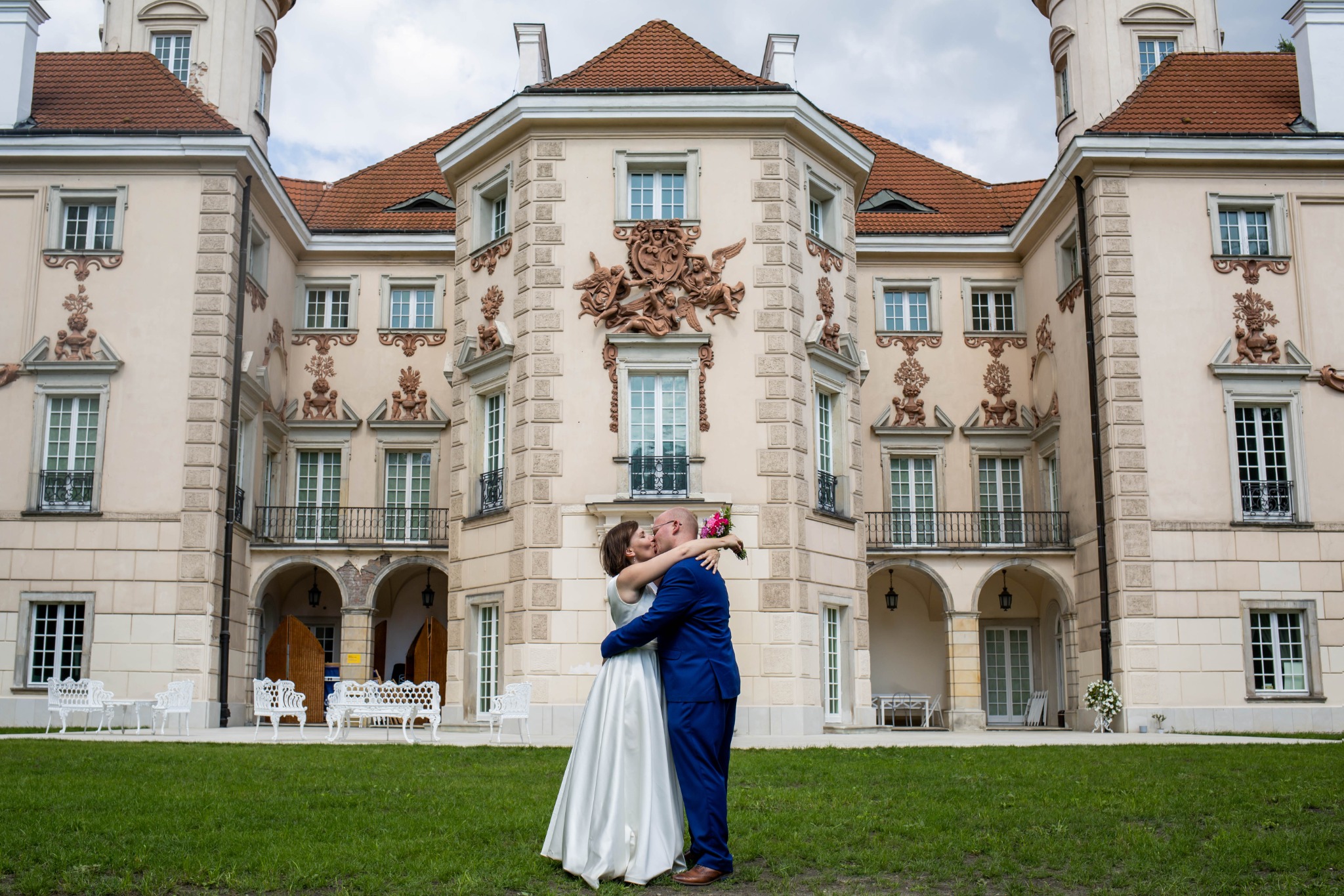 Pocałunek pary młodej na tle jasnego pałacu z dekoracyjnymi płaskorzeźbami i czerwoną dachówką.