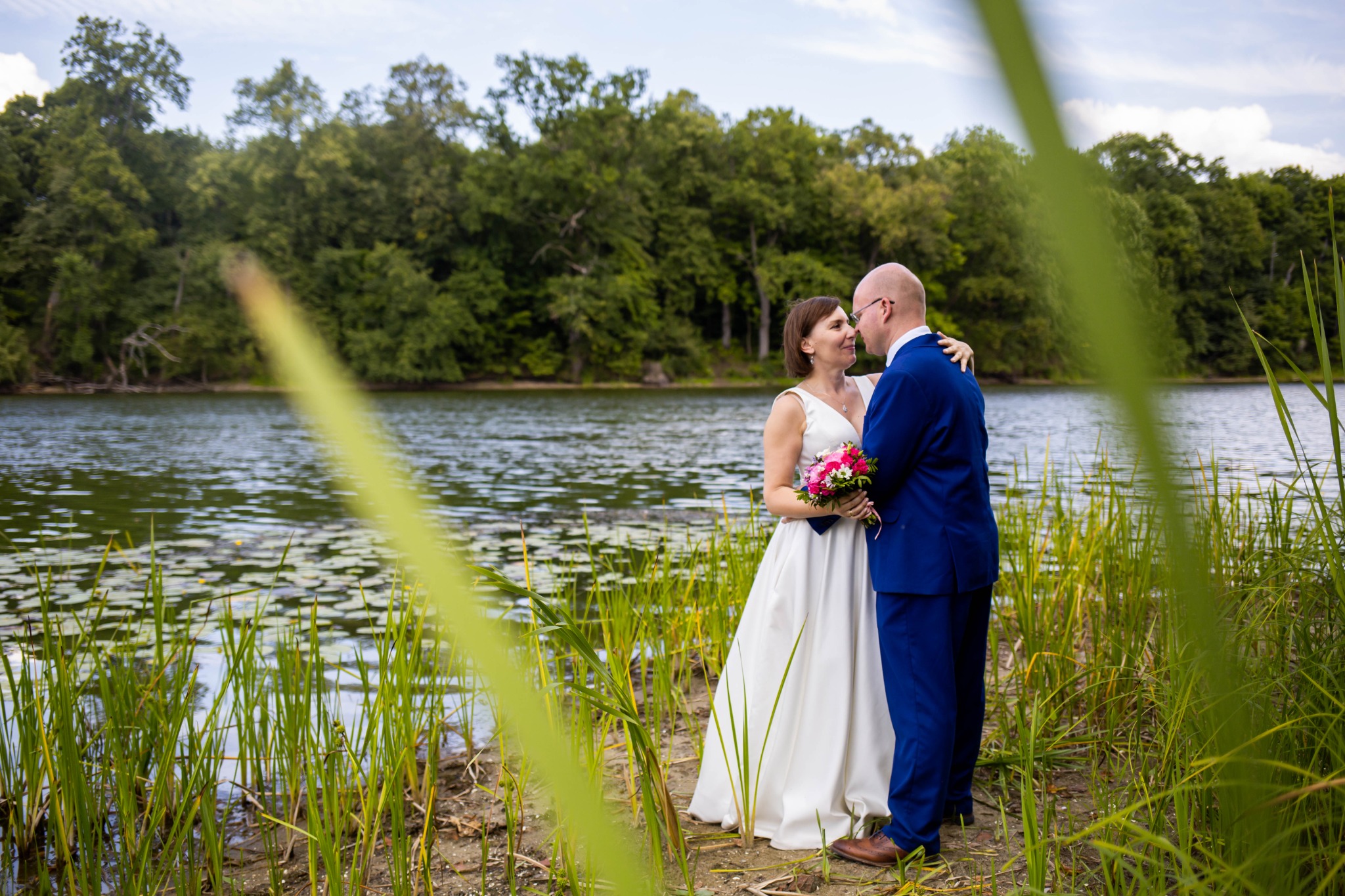 Para młoda w objęciach na brzegu jeziora, panna młoda w białej sukni trzyma bukiet różowych kwiatów, pan młody w niebieskim garniturze, w tle woda i zielony las.