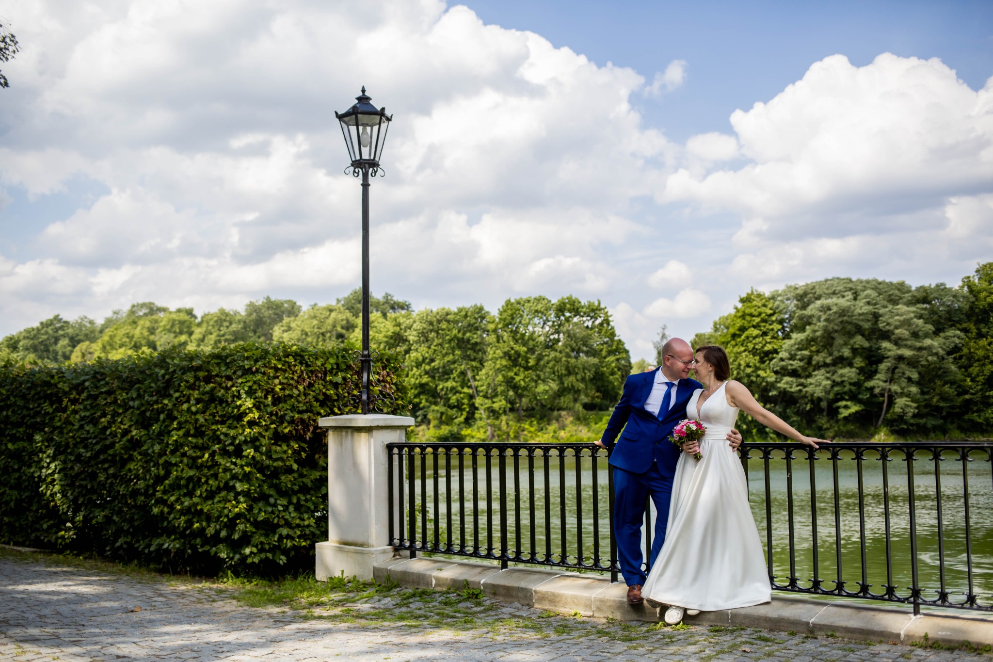 Para młoda w objęciach na tle jeziora, parkowej latarni i zieleni, on w niebieskim garniturze, ona w białej sukni ślubnej z bukietem.