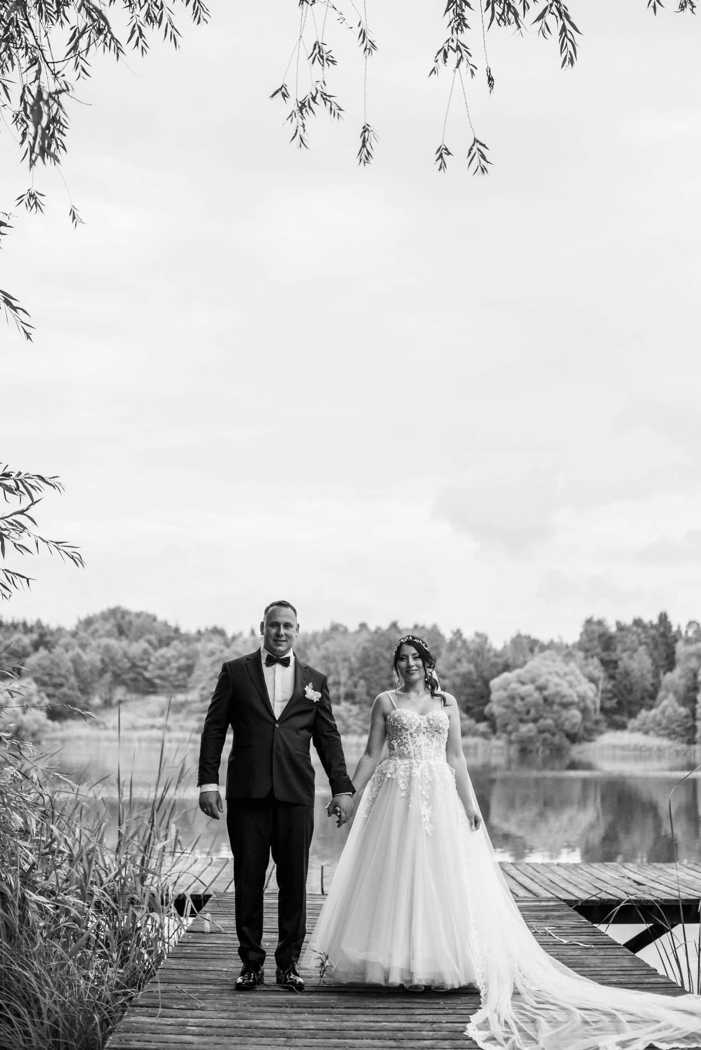 Para młoda trzymająca się za ręce na drewnianym pomoście nad jeziorem, czarno-białe zdjęcie z wiszącymi gałązkami drzew nad głowami.