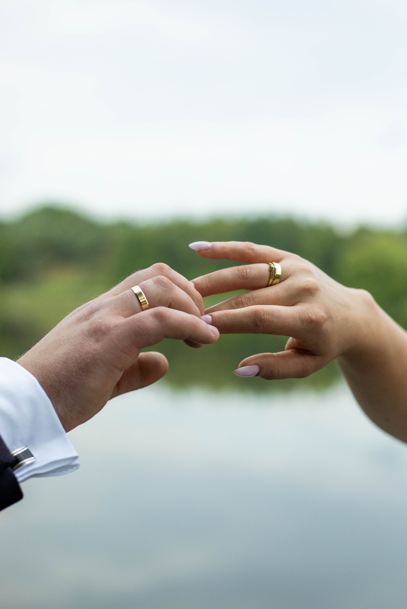 Zbliżenie dłoni pary młodej z złotymi obrączkami na tle jeziora i zieleni, mankiet koszuli męskiej z widocznymi spinkami.