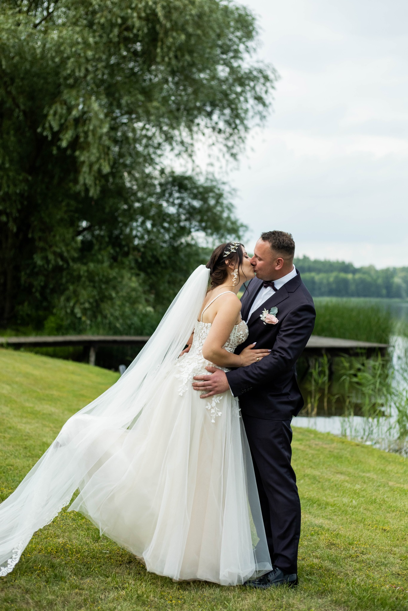 Para młoda w objęciach, pocałunek na tle jeziora i drewnianego pomostu, delikatny wiatr unosi welon panny młodej.