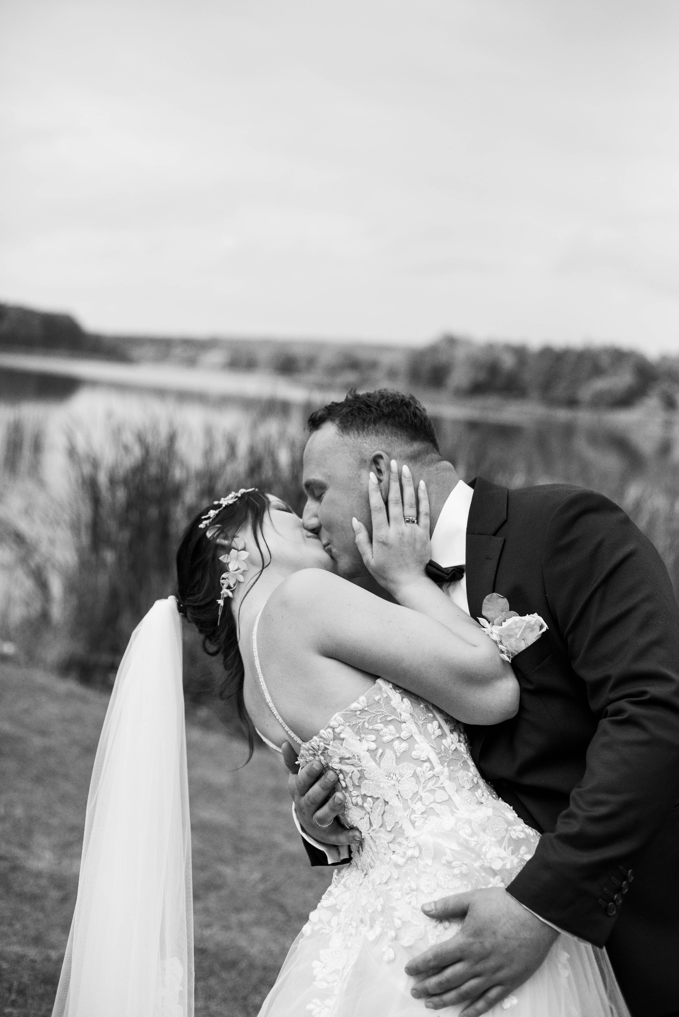 Czarno-białe zdjęcie pocałunku nowożeńców na tle jeziora i trzcin; panna młoda w sukni z koronkowymi aplikacjami, welonie i ozdobie we włosach, pan młody w smokingu z butonierką.