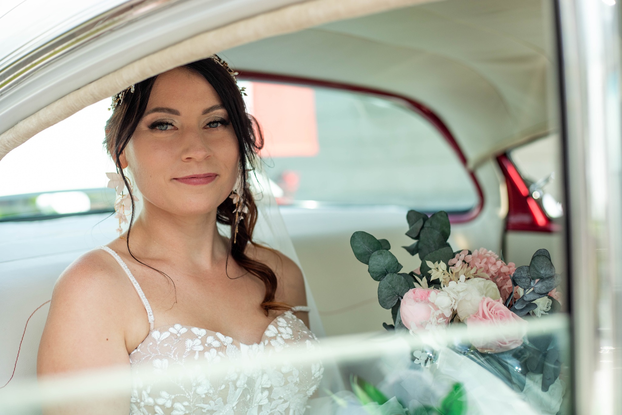 Panna młoda w sukni ślubnej z bukietem kwiatów siedząca w białym samochodzie retro z czerwonymi elementami, widok przez szybę.
