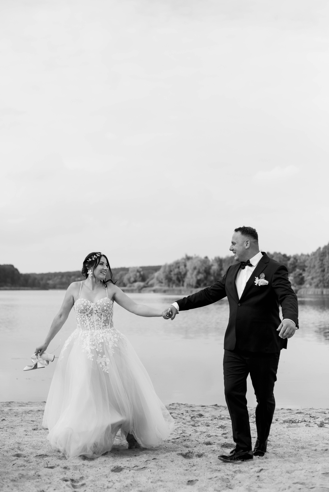 Para młoda w czerni i bieli, trzymająca się za ręce na piaszczystej plaży nad jeziorem. Panna młoda w sukni z koronkowymi aplikacjami trzyma białe buty w dłoni.
