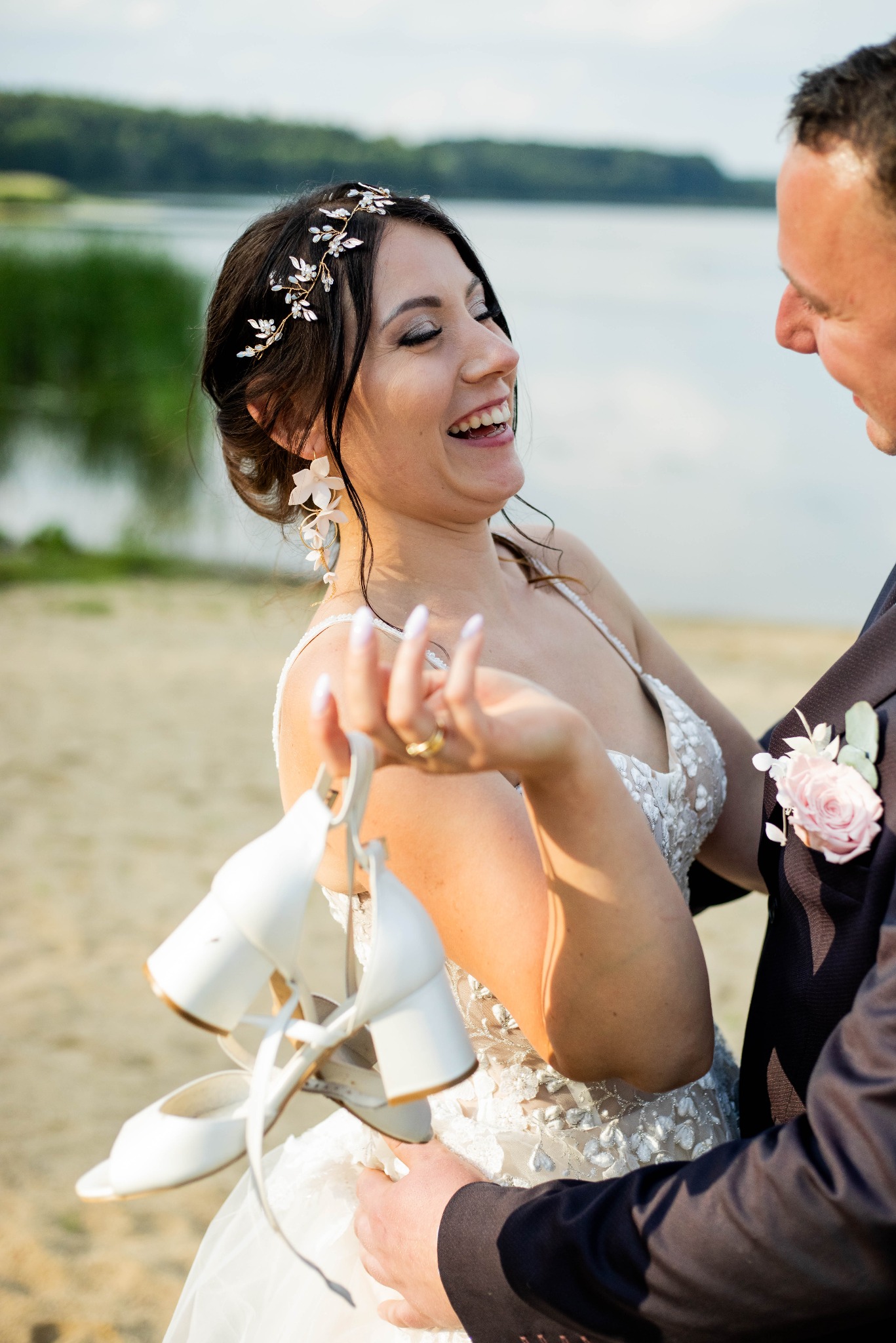 Uśmiechnięta panna młoda w sukni ślubnej z tiulowym dołem, trzymająca białe buty na obcasie na tle jeziora i drzew, obok stoi pan młody w garniturze z butonierką.