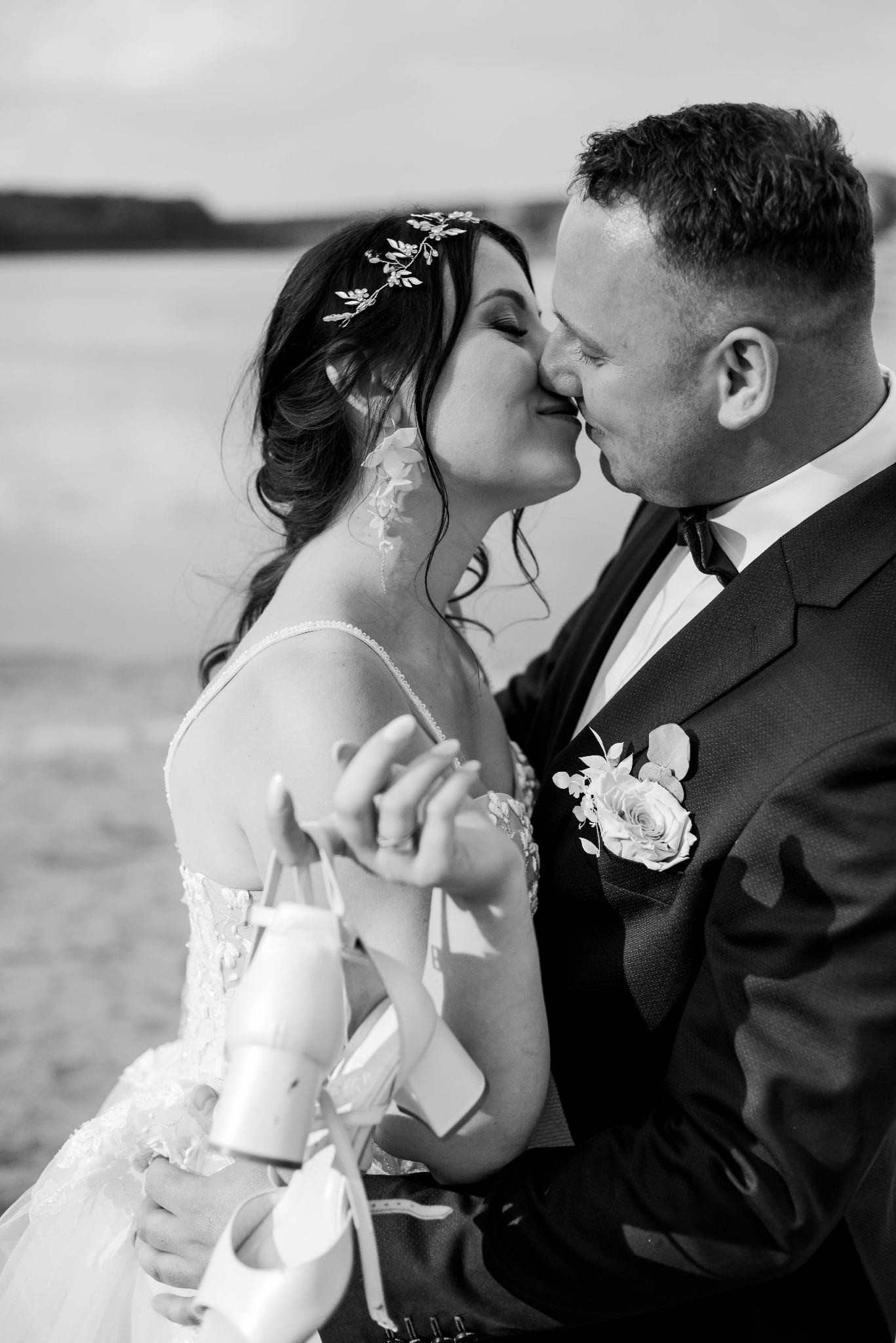 Czarno-białe zbliżenie całującej się pary młodej: panna młoda z ozdobą we włosach i kolczykami w kształcie kwiatów trzyma buty, pan młody w garniturze z butonierką z białej róży.