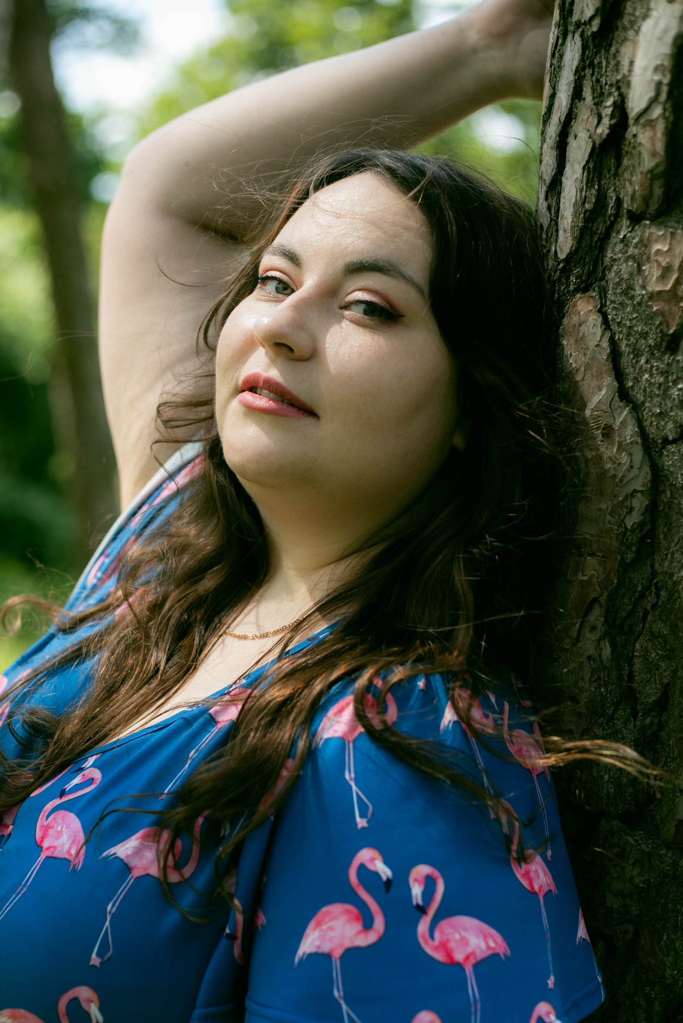 Portret kobiety w niebieskiej sukience z flamingami, opierającej się o korę drzewa, z delikatnym uśmiechem i spojrzeniem w górę.