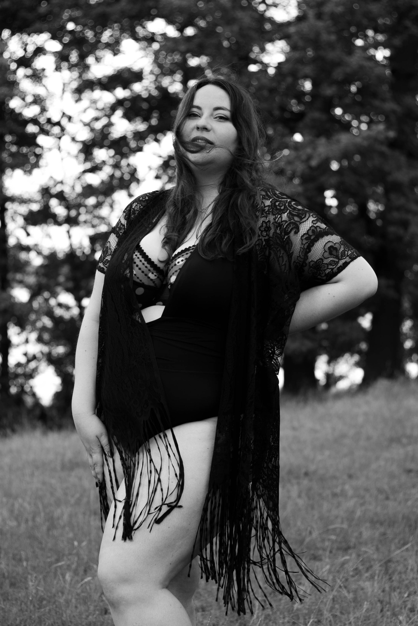 Czarno-białe zdjęcie kobiety w bieliźnie i koronkowym narzutce z frędzlami, stojącej na łące z drzewami w tle, włosy rozwiane przez wiatr.