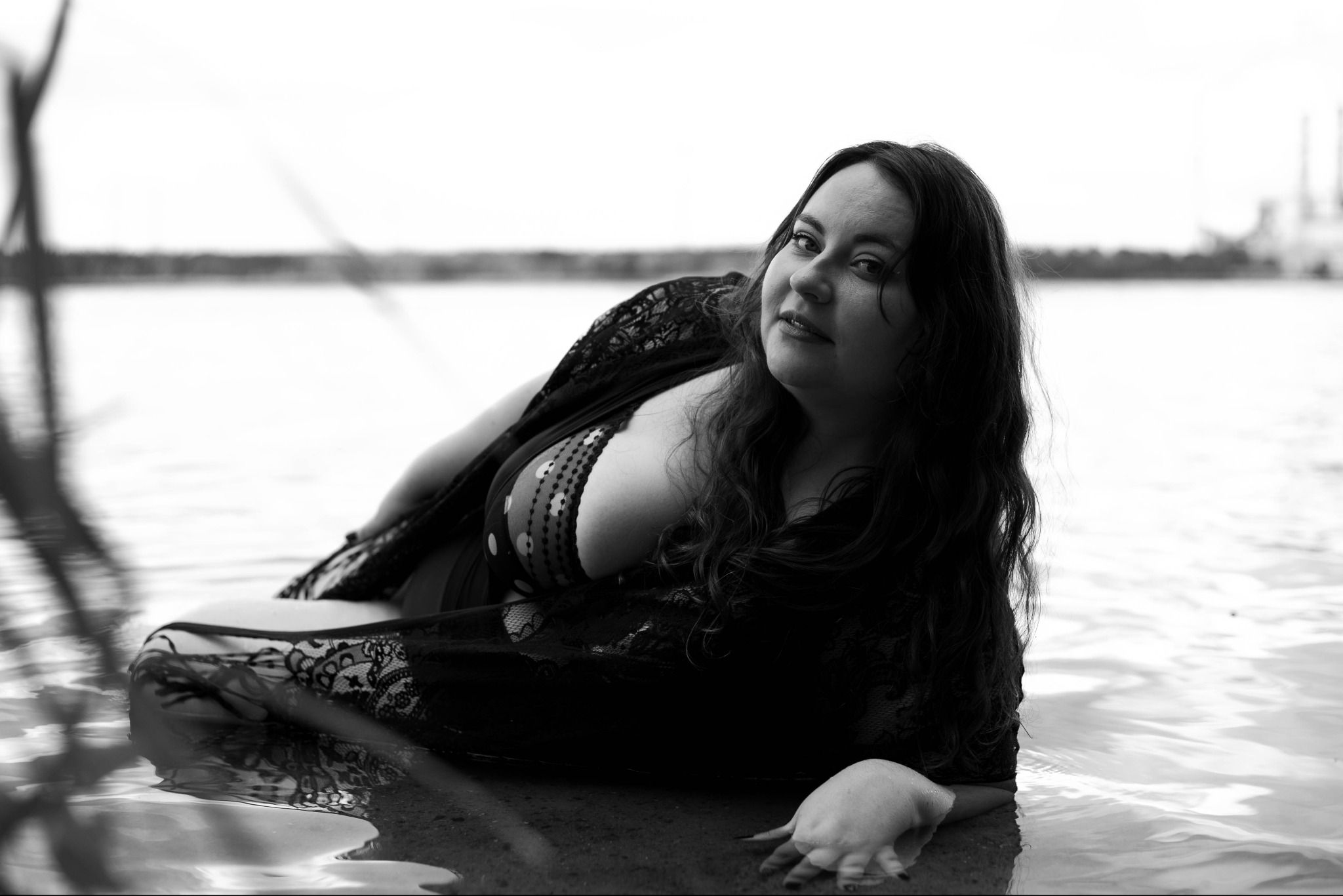 Czarno-biała fotografia kobiety w wodzie, ubrana w koronkowy szal, leżącej na brzegu z fabryką w tle, patrzącej w obiektyw.