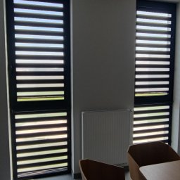 AGAFIRANY Agnieszka Biesiadecka - Dwa okna z czarnymi żaluzjami fasadowymi, częściowo otwarte, w jasnym biurze z grzejnikiem i fragmentem stołu z krzesłami.