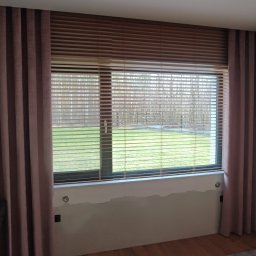 AGAFIRANY Agnieszka Biesiadecka - Drewniane poziome listwy osłaniające okno, uzupełnione zasłonami w odcieniach brudnego różu, częściowo zakrywające okno z widokiem na zieleń i drzewa.