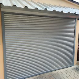 AGAFIRANY Agnieszka Biesiadecka - Zamknięta, szara, metalowa osłona okienna z poziomymi lamelami, zamontowana na budynku o beżowej elewacji, widoczna z poziomu chodnika wyłożonego kostką.