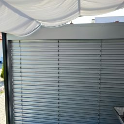 AGAFIRANY Agnieszka Biesiadecka - Zewnętrzne metalowe lamele osłonowe na tarasie z widokiem na ogród i fragment domu, widoczny grill z boku.