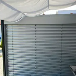 Zewnętrzne metalowe lamele osłonowe na tarasie z widokiem na ogród i fragment domu, widoczny grill z boku.