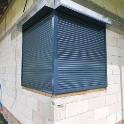 AGAFIRANY Agnieszka Biesiadecka - Zamknięta, grafitowa osłona okienna na elewacji z białego gazobetonu, widok z zewnątrz w trakcie prac wykończeniowych.