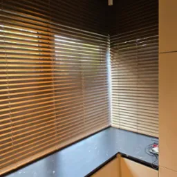 Zaciemnione brązowe poziome listwy osłaniające okna w rogu pokoju, z widocznym fragmentem blatu i szafek, oświetlenie górne.