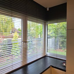 AGAFIRANY Agnieszka Biesiadecka - Wnętrze pokoju z widokiem na zieleń przez okno z poziomymi żaluzjami w kolorze jasnego drewna, czarny blat pod oknem i fragment zabudowy kuchennej.