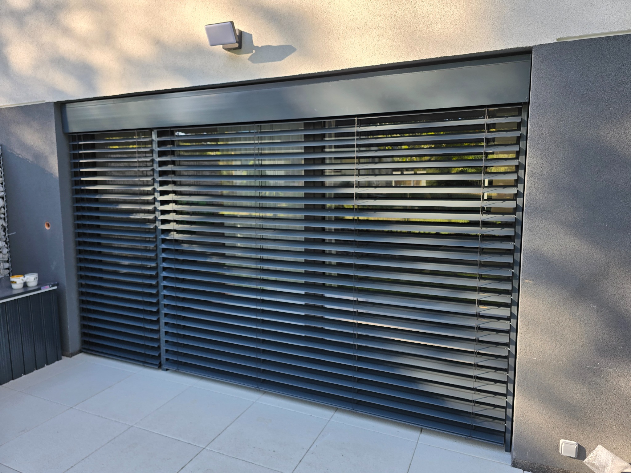 Szare poziome osłony okienne zamontowane na tarasie, widok z zewnątrz, z widocznymi elementami konstrukcyjnymi i fragmentem elewacji budynku.