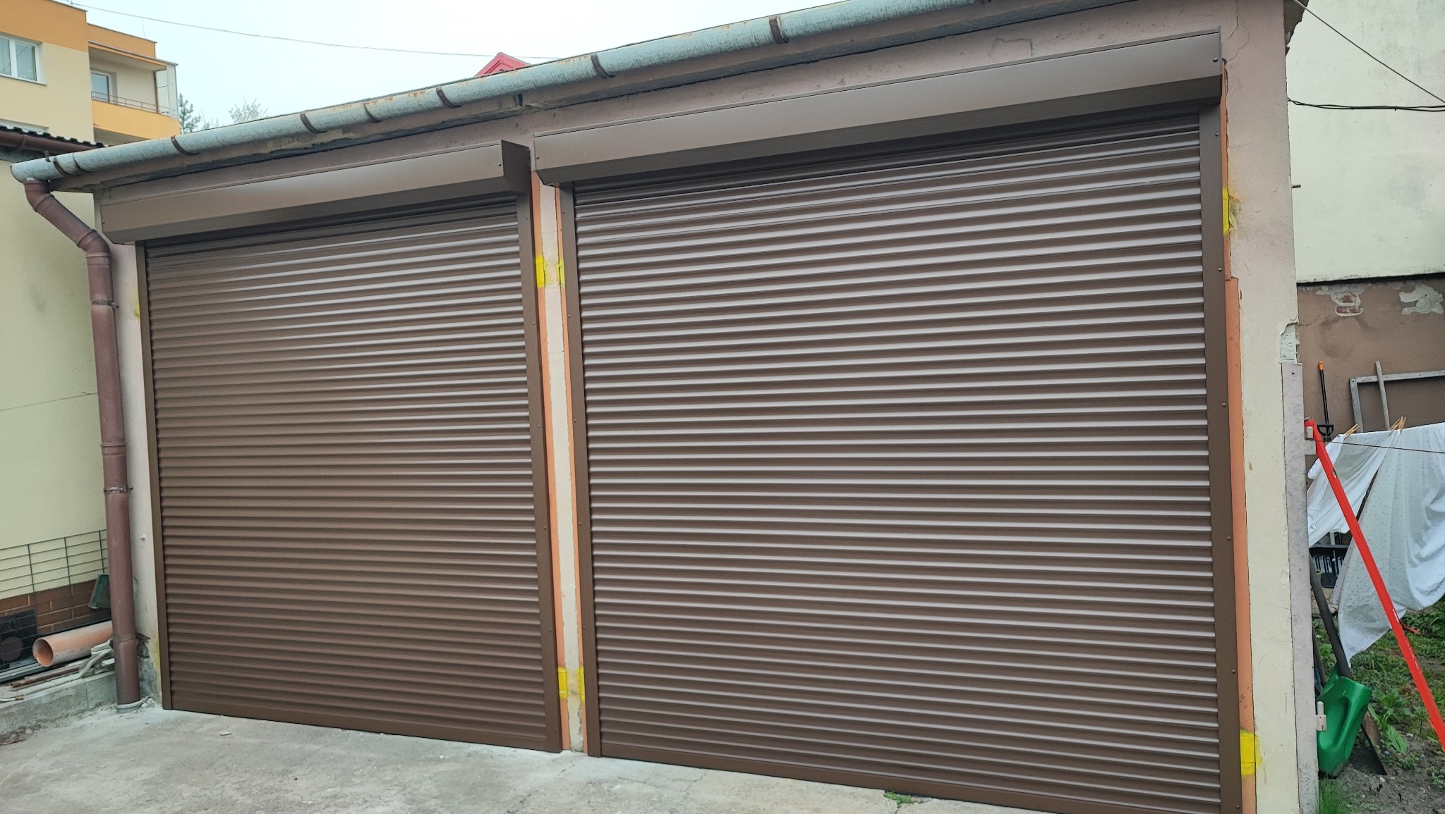 Dwa garaże z brązowymi, zamkniętymi osłonami okiennymi, z widocznymi elementami montażowymi i niedokończonymi pracami wykończeniowymi ścian.