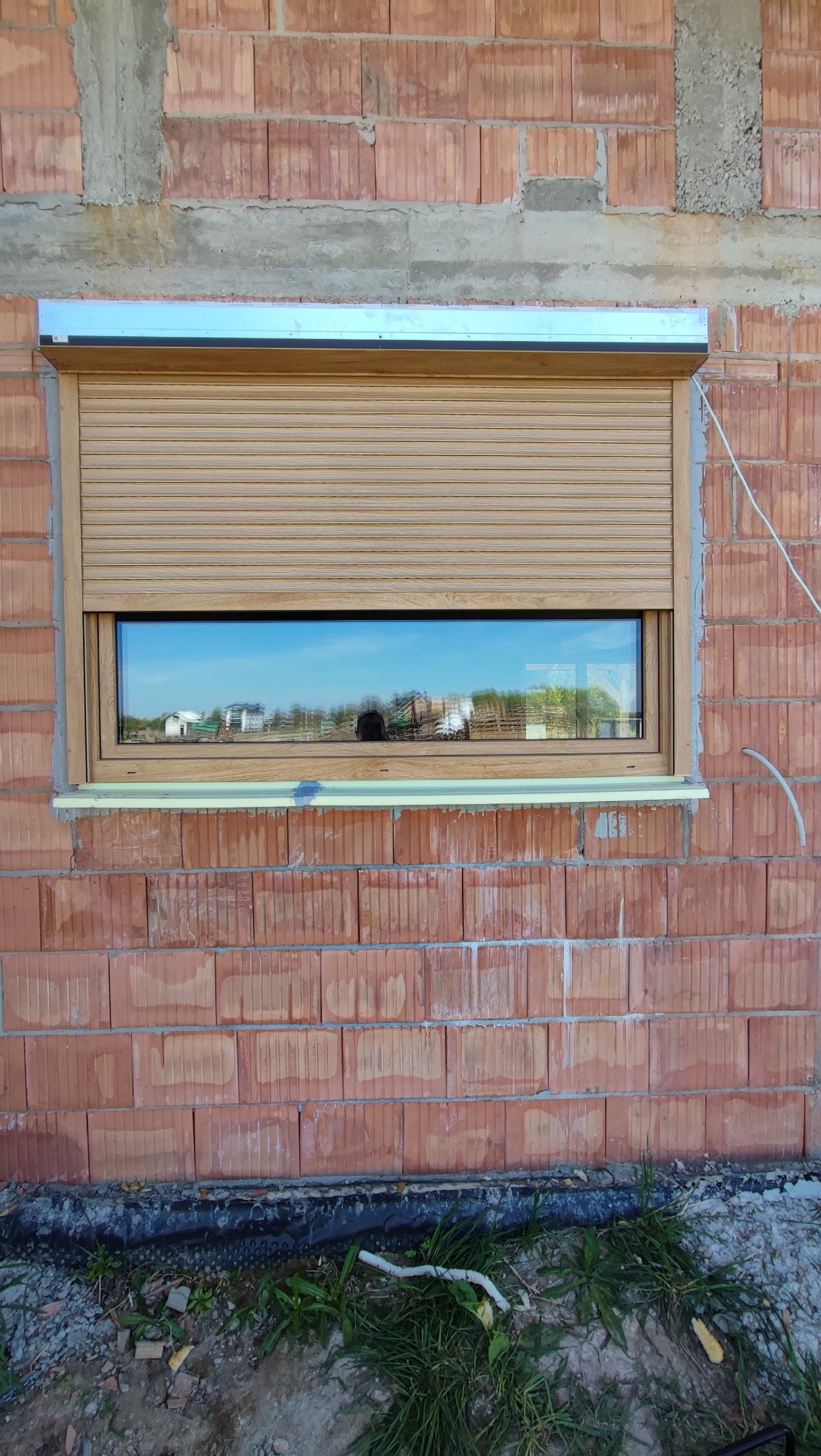 Zamknięta, jasnobrązowa osłona okienna zamontowana w surowej, ceglanej ścianie budynku w trakcie budowy, z widocznym fragmentem nieotynkowanego nadproża.
