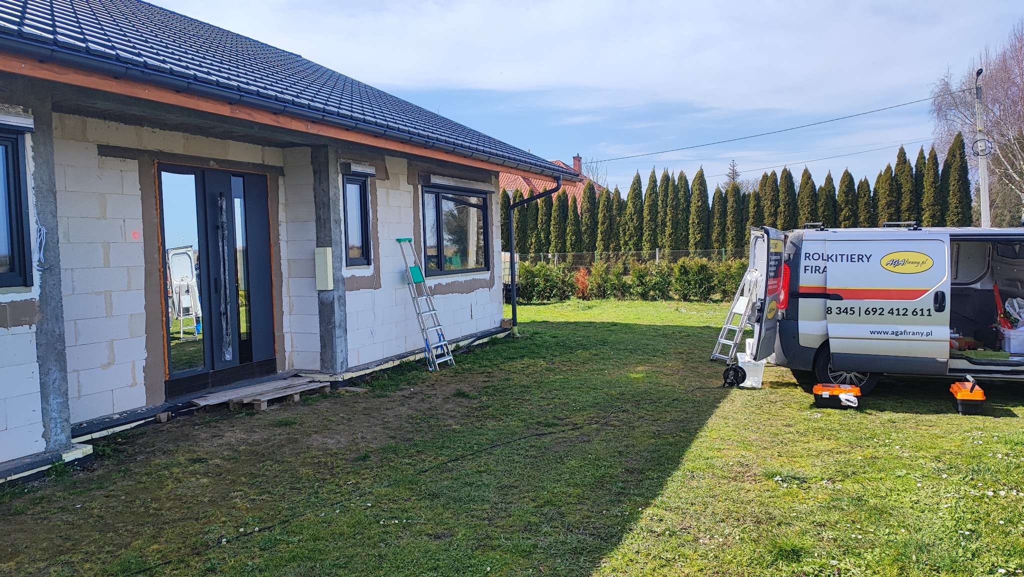 Montaż stolarki okiennej i drzwiowej w nowo budowanym domu jednorodzinnym, widoczny samochód dostawczy z reklamą firmy monterskiej oraz drabiny oparta o ścianę budynku.