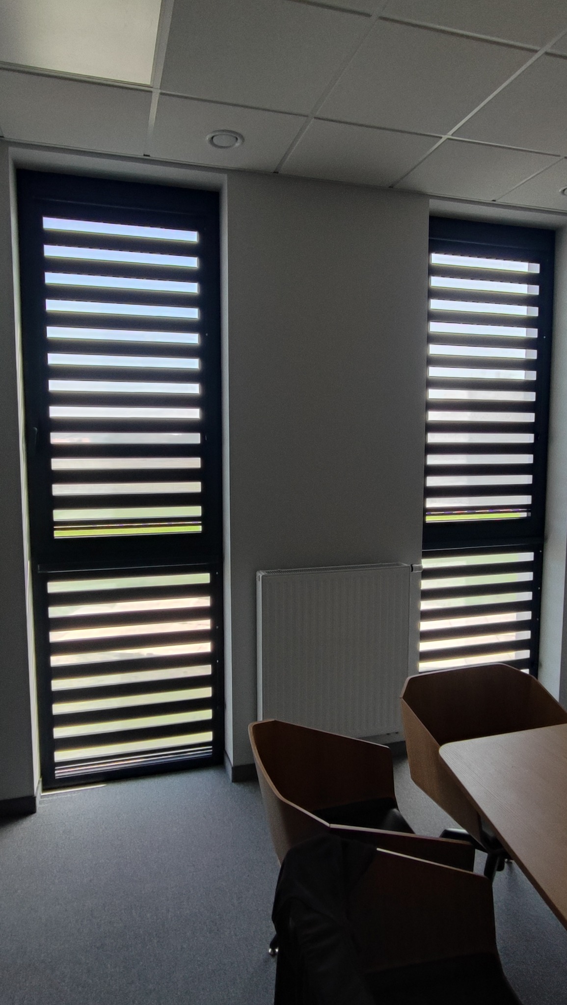 Dwa okna z czarnymi żaluzjami fasadowymi, częściowo otwarte, w jasnym biurze z grzejnikiem i fragmentem stołu z krzesłami.