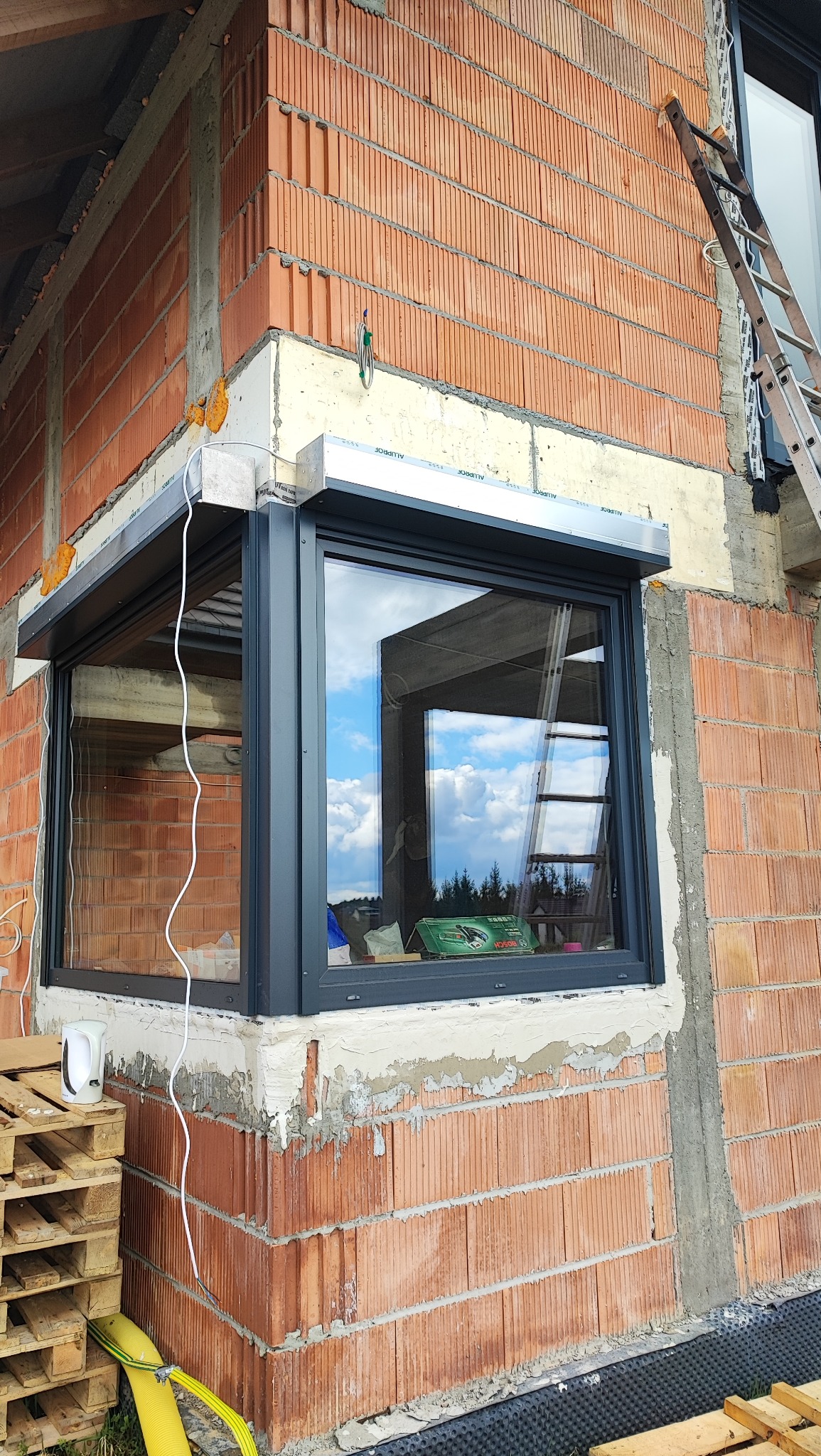 Montaż szarej rolety zewnętrznej na oknie w surowym budynku z czerwonej cegły, widoczna drabina oparta o ścianę i paleta z materiałami budowlanymi u dołu.
