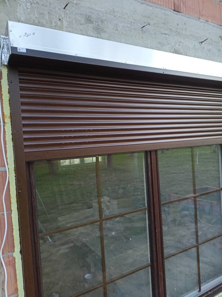 Ciemnobrązowa, częściowo opuszczona osłona okienna z aluminiową obudową, zamontowana na oknie z ciemną ramą, widoczna na tle surowego muru z wystającymi elementami montażowymi.