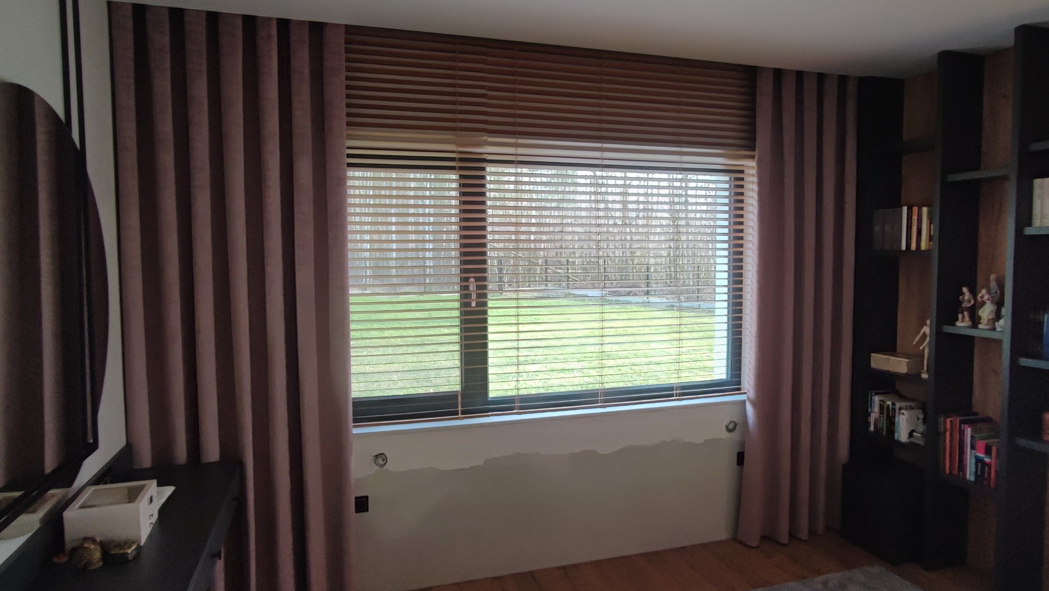 Drewniane poziome listwy osłaniające okno, uzupełnione zasłonami w odcieniach brudnego różu, częściowo zakrywające okno z widokiem na zieleń i drzewa.