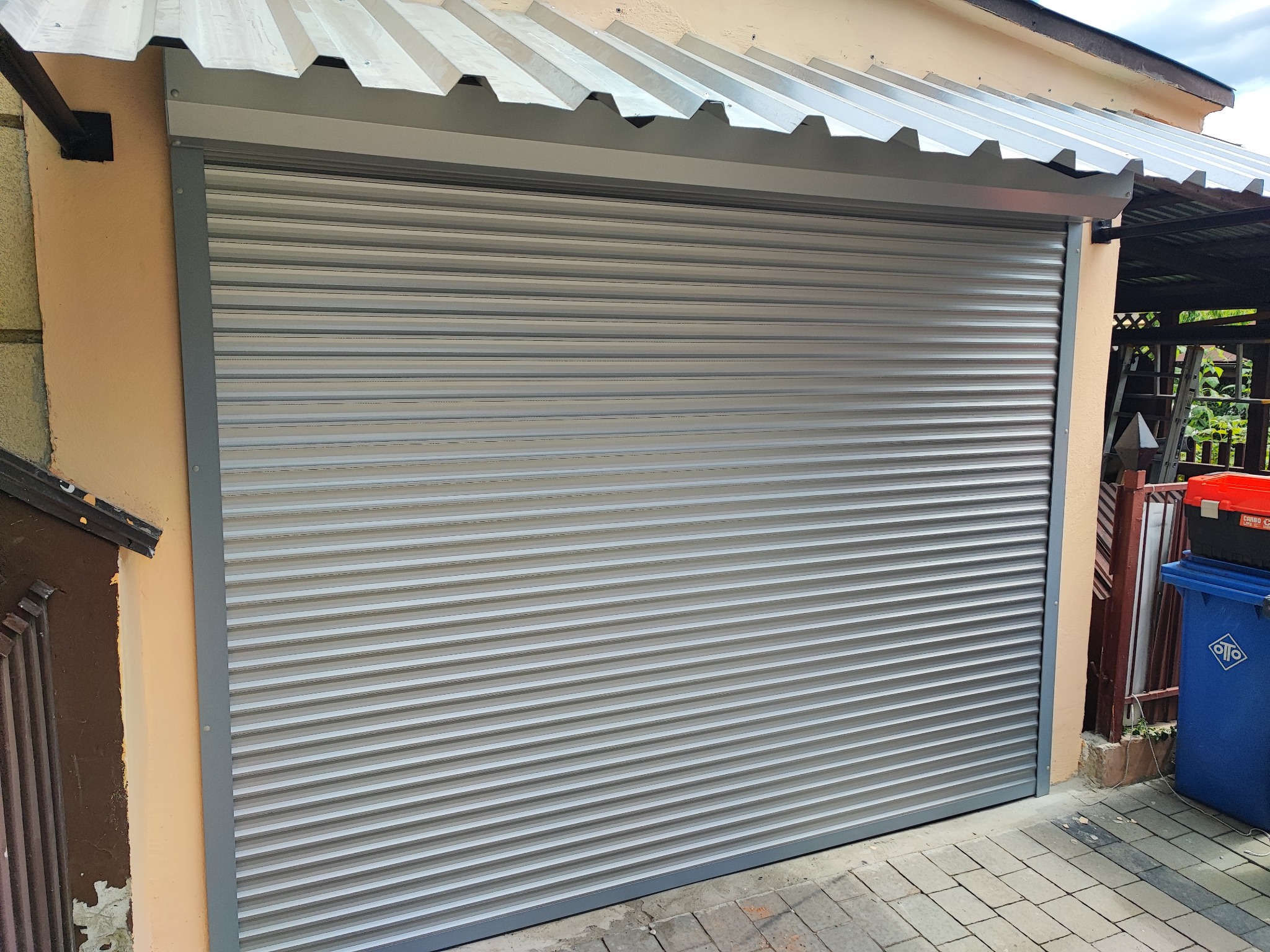 Zamknięta, szara, metalowa osłona okienna z poziomymi lamelami, zamontowana na budynku o beżowej elewacji, widoczna z poziomu chodnika wyłożonego kostką.