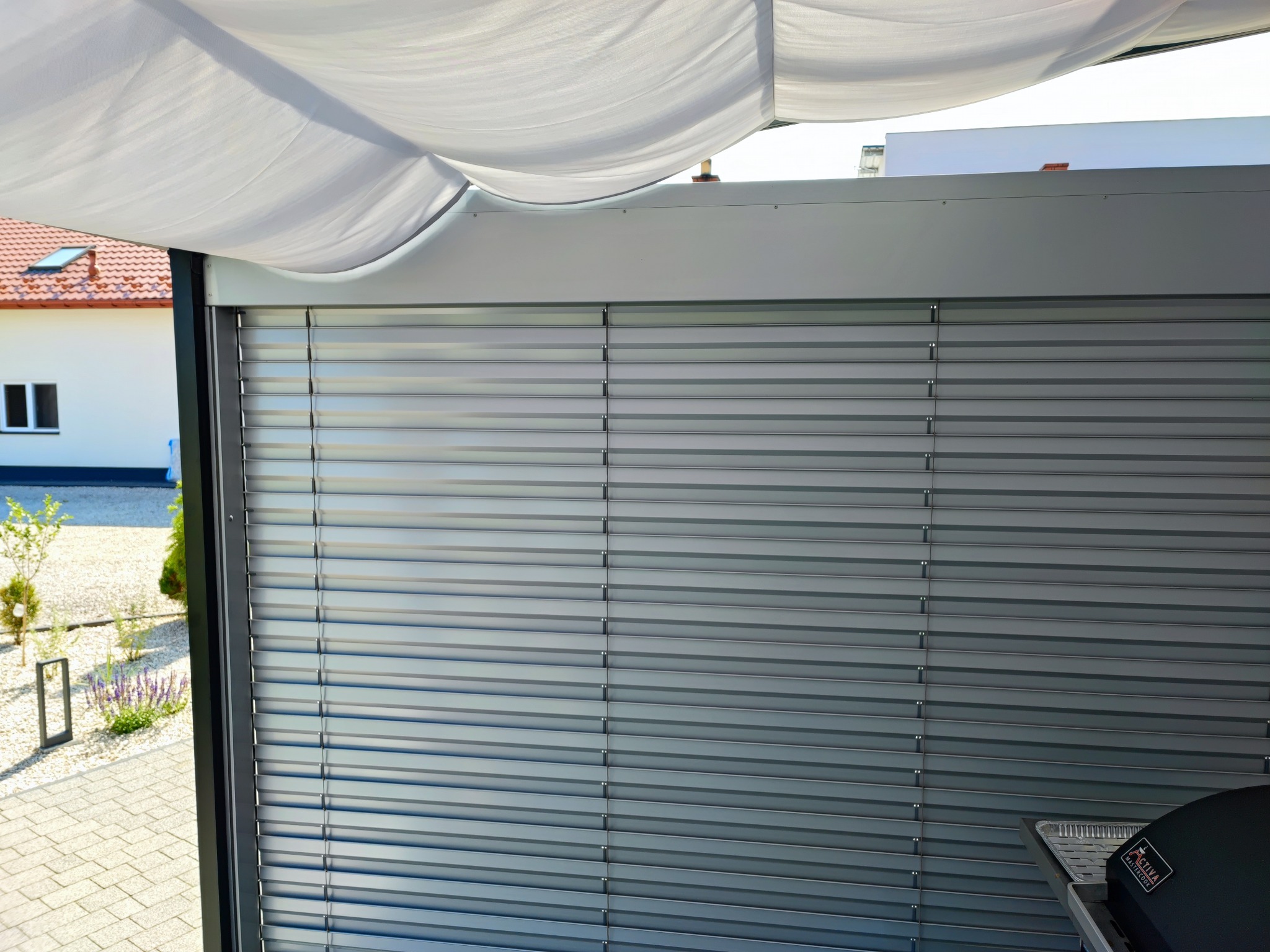 Zewnętrzne metalowe lamele osłonowe na tarasie z widokiem na ogród i fragment domu, widoczny grill z boku.