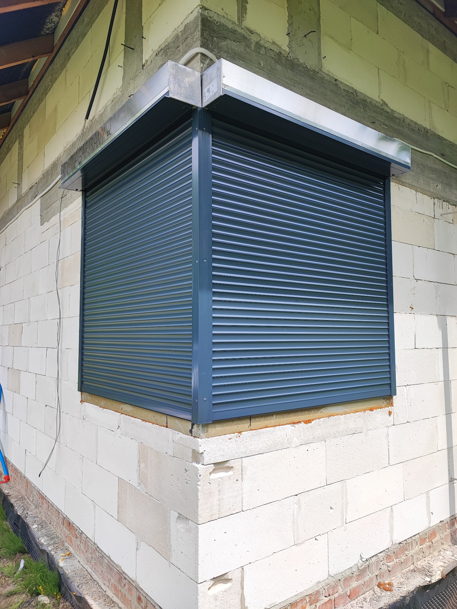 Zamknięta, grafitowa osłona okienna na elewacji z białego gazobetonu, widok z zewnątrz w trakcie prac wykończeniowych.