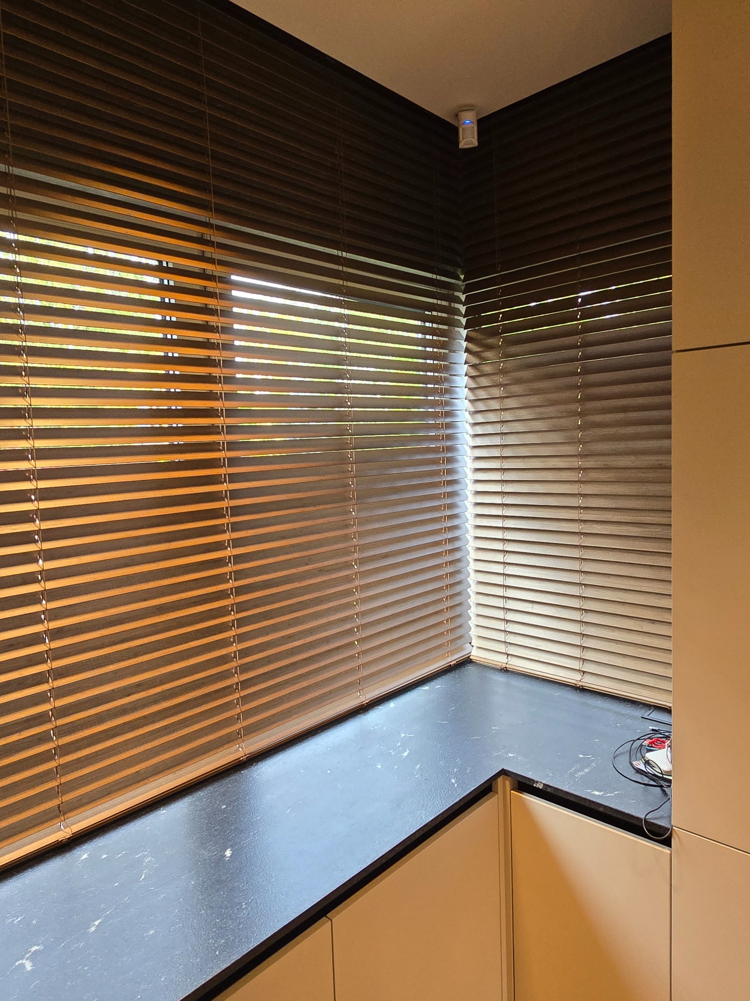 Zaciemnione brązowe poziome listwy osłaniające okna w rogu pokoju, z widocznym fragmentem blatu i szafek, oświetlenie górne.