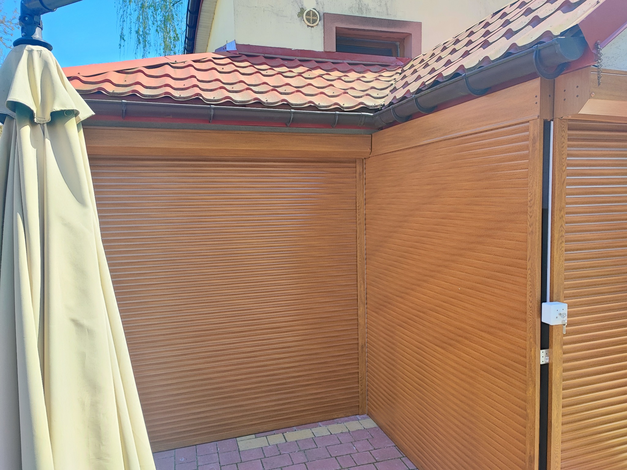 Zewnętrzna konstrukcja z brązowych paneli z poziomymi żłobieniami, przypominająca zamkniętą osłonę, z widocznym fragmentem zwiniętego parasola przeciwsłonecznego po lewej stronie, na tle kostki...