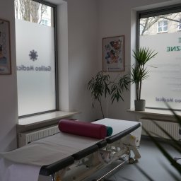 Galileo Medical - Fizjoterapia, Rehabilitacja, Masaże - Warszawa Mokotów - Jasne wnętrze gabinetu z kozetką przykrytą białym prześcieradłem, różowym wałkiem i zielonym półwałkiem, dekorowane roślinami doniczkowymi i plakatem na ścianie, okno z napisem Galileo Medical.