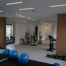 Galileo Medical - Fizjoterapia, Rehabilitacja, Masaże - Warszawa Mokotów - Jasne, minimalistyczne wnętrze siłowni wyposażone w niebieskie piłki gimnastyczne, kettlebells, rower stacjonarny, drabinki gimnastyczne i rośliny doniczkowe, odbijające się w dużych lustrach.