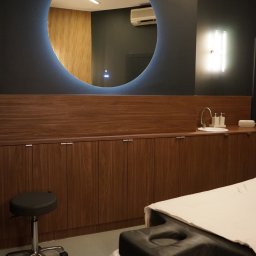 Galileo Medical - Fizjoterapia, Rehabilitacja, Masaże - Warszawa Mokotów - Nowoczesne wnętrze gabinetu z okrągłym lustrem podświetlonym na niebiesko, drewnianymi szafkami, umywalką i leżanką z białym prześcieradłem, oświetlone lampą ścienną.