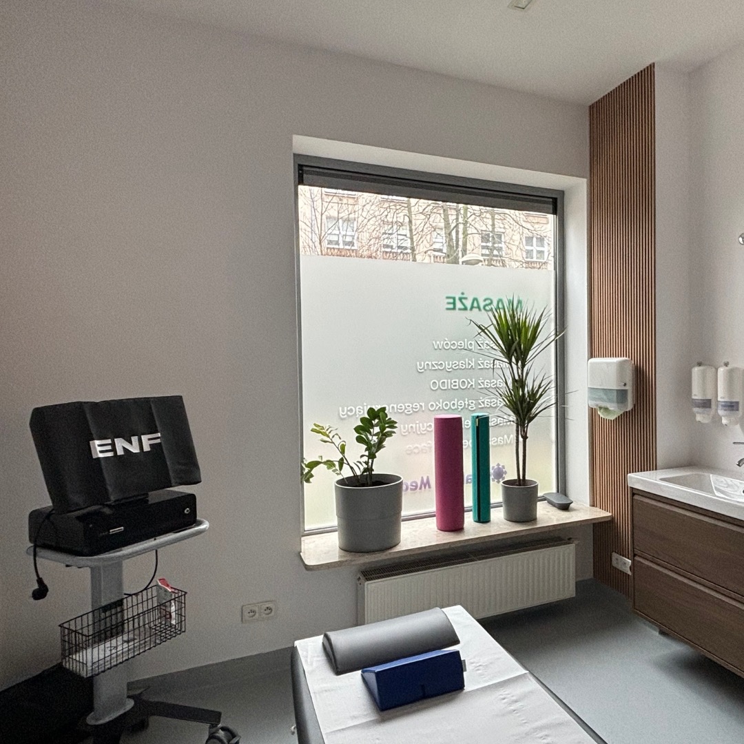 Jasne wnętrze gabinetu z łóżkiem do zabiegów, urządzeniem ENF na wózku, oknem z napisem 'MASAŻE', roślinami w doniczkach i drewnianym panelem ściennym.