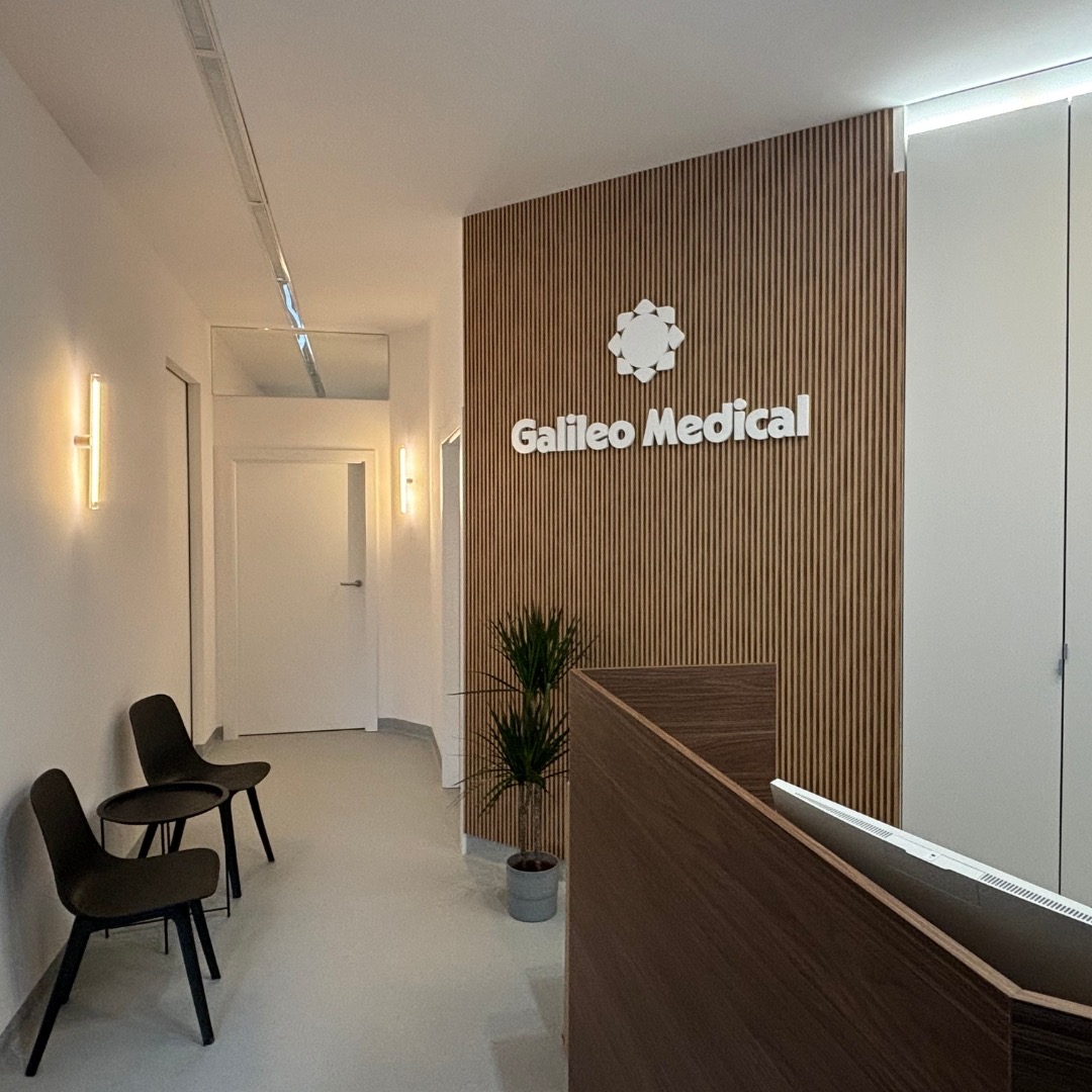 Wnętrze poczekalni kliniki medycznej z recepcją, ścianą z drewnianych paneli, logo 'Galileo Medical', rośliną w doniczce i krzesłami dla pacjentów.