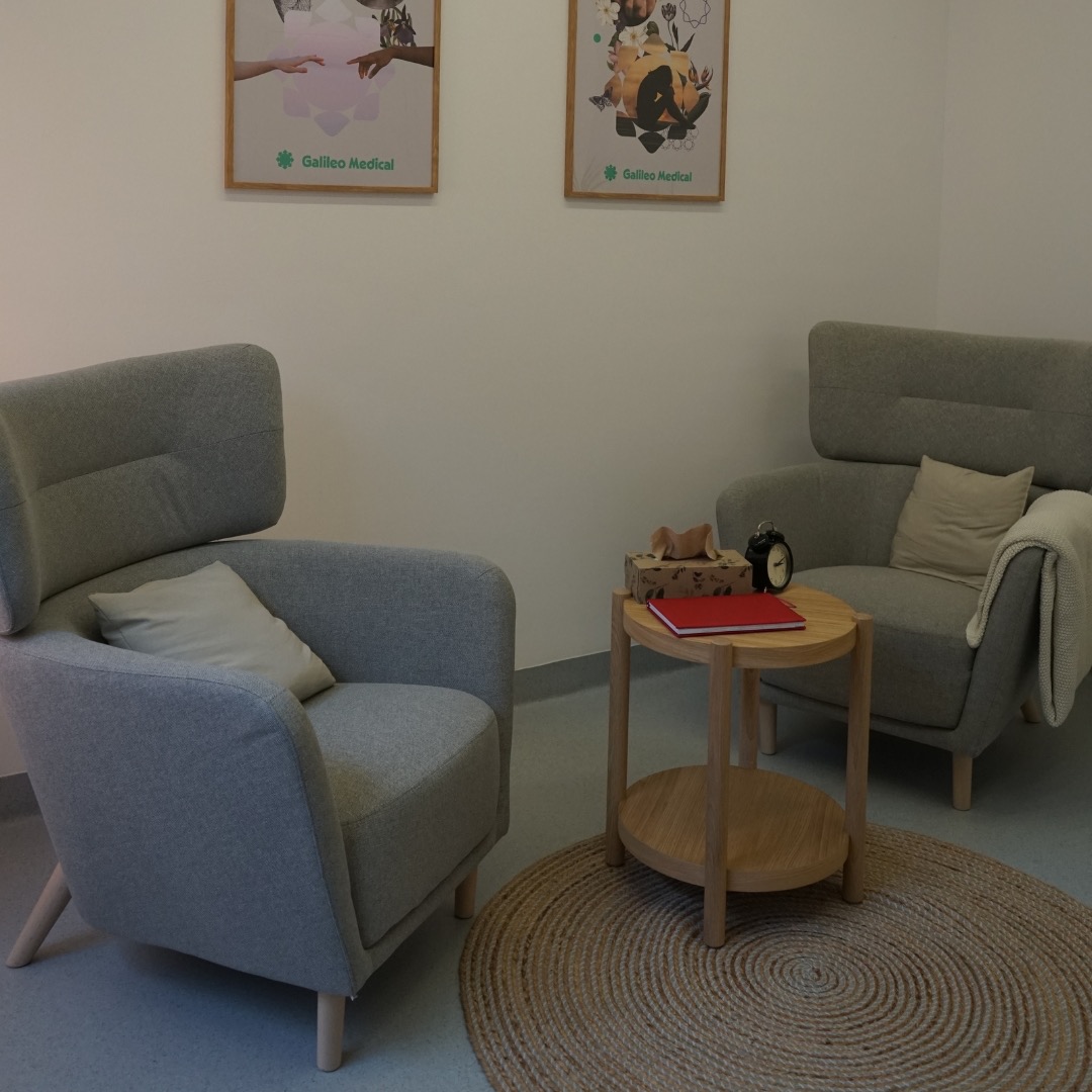 Jasne, minimalistyczne wnętrze gabinetu z dwoma szarymi fotelami, okrągłym drewnianym stolikiem z czerwoną książką i zegarem, oraz dwoma obrazami z logo Galileo Medical na ścianie.