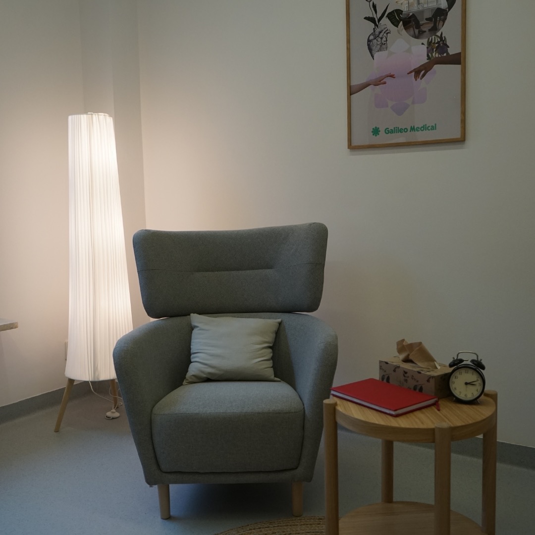 Jasny gabinet z szarym fotelem, poduszką, lampą podłogową, stolikiem z czerwoną książką, chusteczkami i zegarem, oraz plakatem 'Galileo Medical' na ścianie.