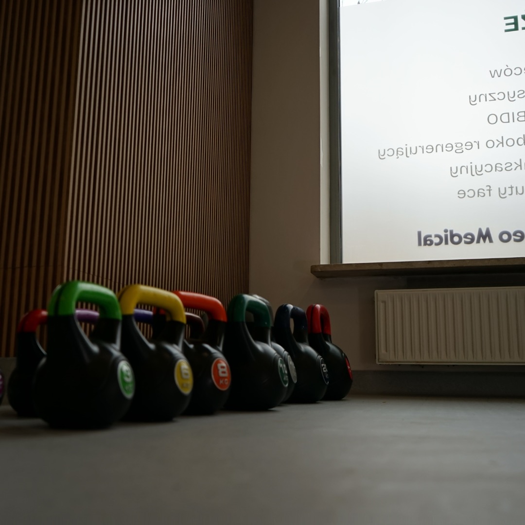 Rząd czarnych kettlebell z kolorowymi uchwytami (zielony, żółty, pomarańczowy, ciemnozielony, fioletowy, czerwony) ułożonych na podłodze w siłowni, widoczna ściana z drewnianych paneli i okno...