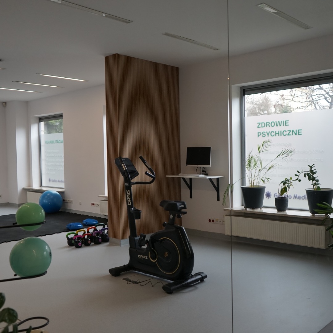 Wnętrze gabinetu z rowerem stacjonarnym, piłkami do ćwiczeń i hantlami kettlebell, okno z napisem 'Zdrowie Psychiczne' i roślinami w doniczkach.