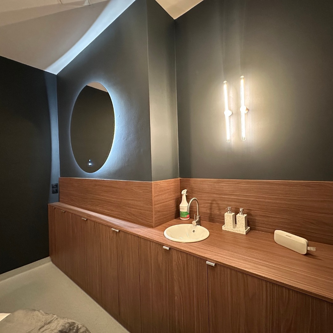 Nowoczesna łazienka z owalnym lustrem podświetlanym od tyłu, minimalistyczną armaturą i drewnianymi szafkami, dwie pionowe lampy na ciemnej ścianie.
