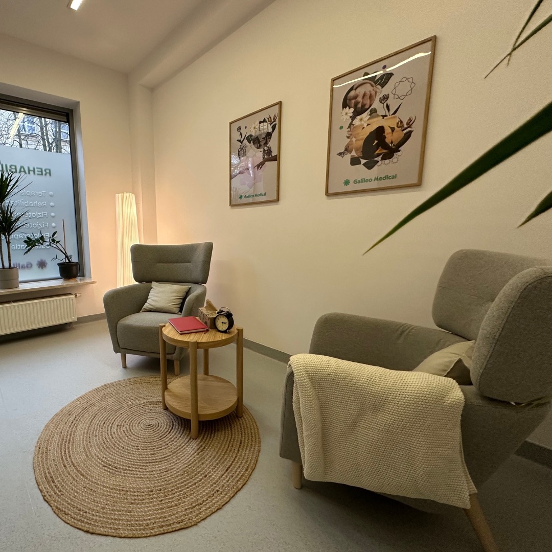 Jasne wnętrze gabinetu z dwoma szarymi fotelami, okrągłym dywanem z juty, małym stolikiem z książką i zegarem, oraz plakatami na ścianie z logo 'Gallico Medical'.