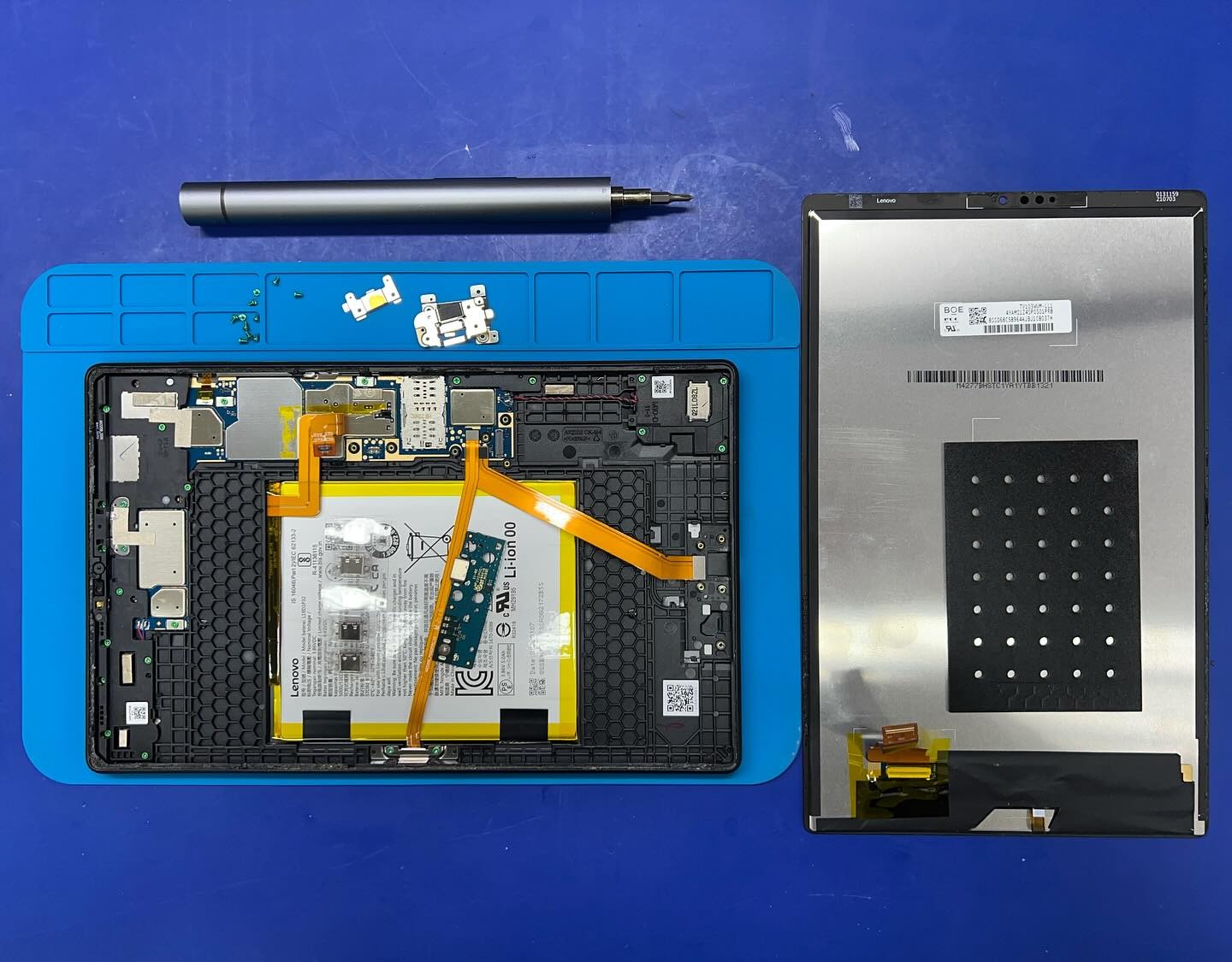 Rozłożony tablet Lenovo na niebieskiej macie serwisowej, widoczne wnętrze z baterią, odłączony ekran, precyzyjny śrubokręt i drobne elementy elektroniczne obok.