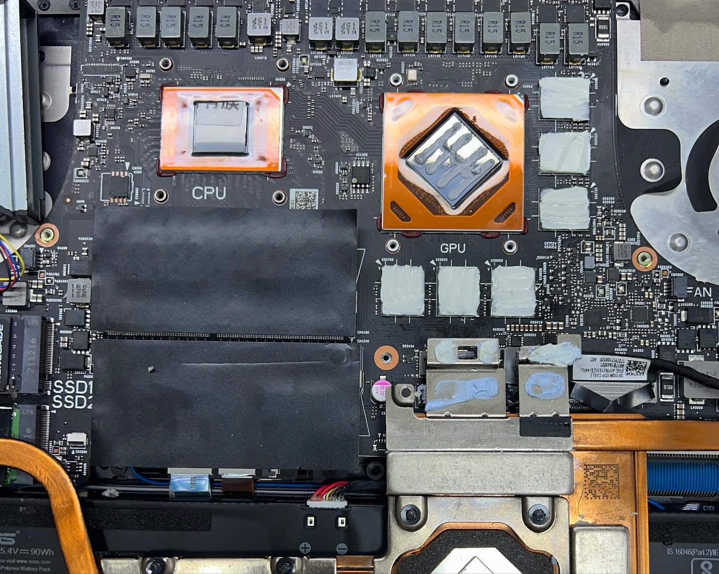 Wnętrze laptopa z widocznymi elementami płyty głównej: procesor CPU z nałożoną pastą termoprzewodzącą, GPU z systemem chłodzenia i termopadami, dyski SSD oraz wentylator. Widoczne liczne układy...