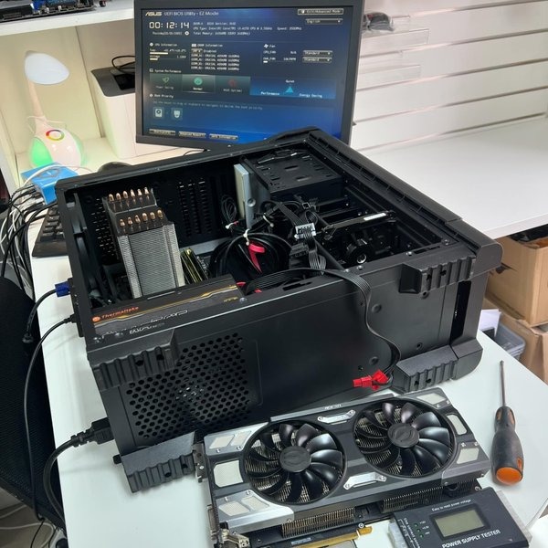Otwarta obudowa komputera stacjonarnego z widocznym chłodzeniem procesora, kartą graficzną Thermaltake, oraz dodatkowymi kartami graficznymi EVGA i miernikiem napięcia zasilacza obok, na biurku...