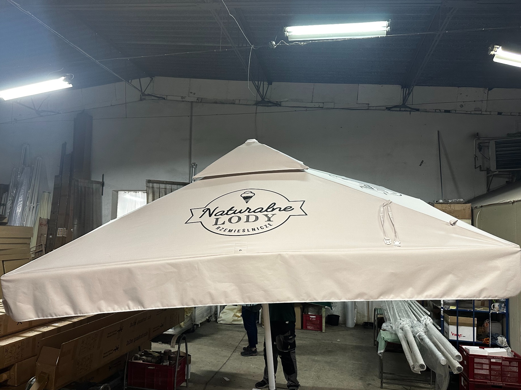 Duży, beżowy namiot z nadrukiem 'Naturalne Lody Rzemieślnicze' w magazynie, widoczne metalowe ramy i elementy konstrukcyjne.